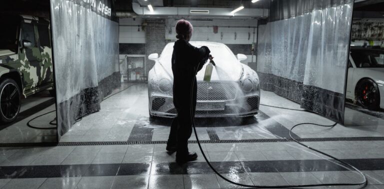 A person using a hose to spray a car with snow foam in an indoor carwash setting.
